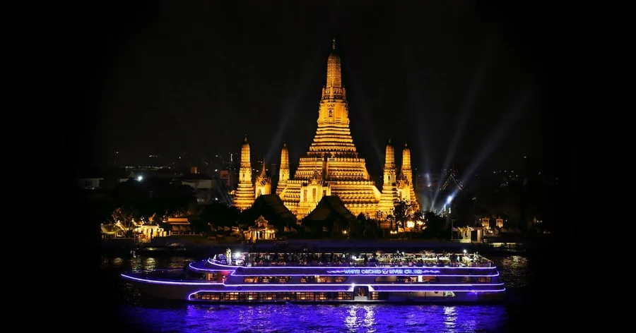 วัดอรุณเปิดไฟสีทองยามค่ำคืนเมื่อมองจากฝั่งตรงข้าม มีเรือล่องแม่น้ำเปิดไฟแล่นผ่านด้านหน้า